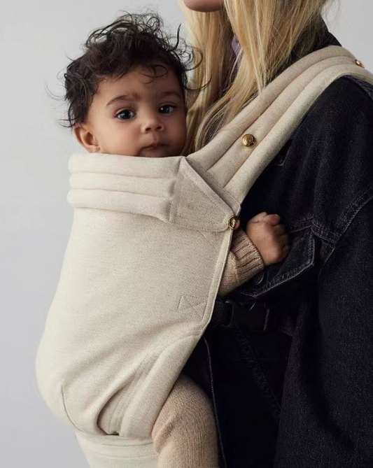 Baby wrapped in a beige carrier held by a person against a plain background